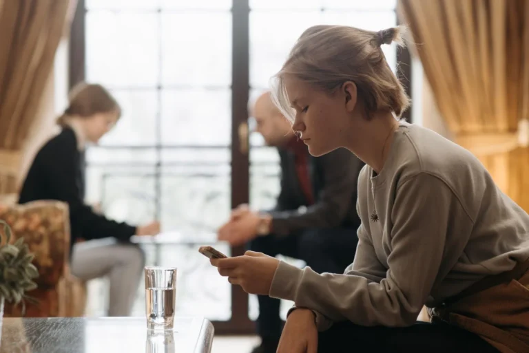 A teenager focuses on a smartphone indoors while adults talk, blurred, in the background.